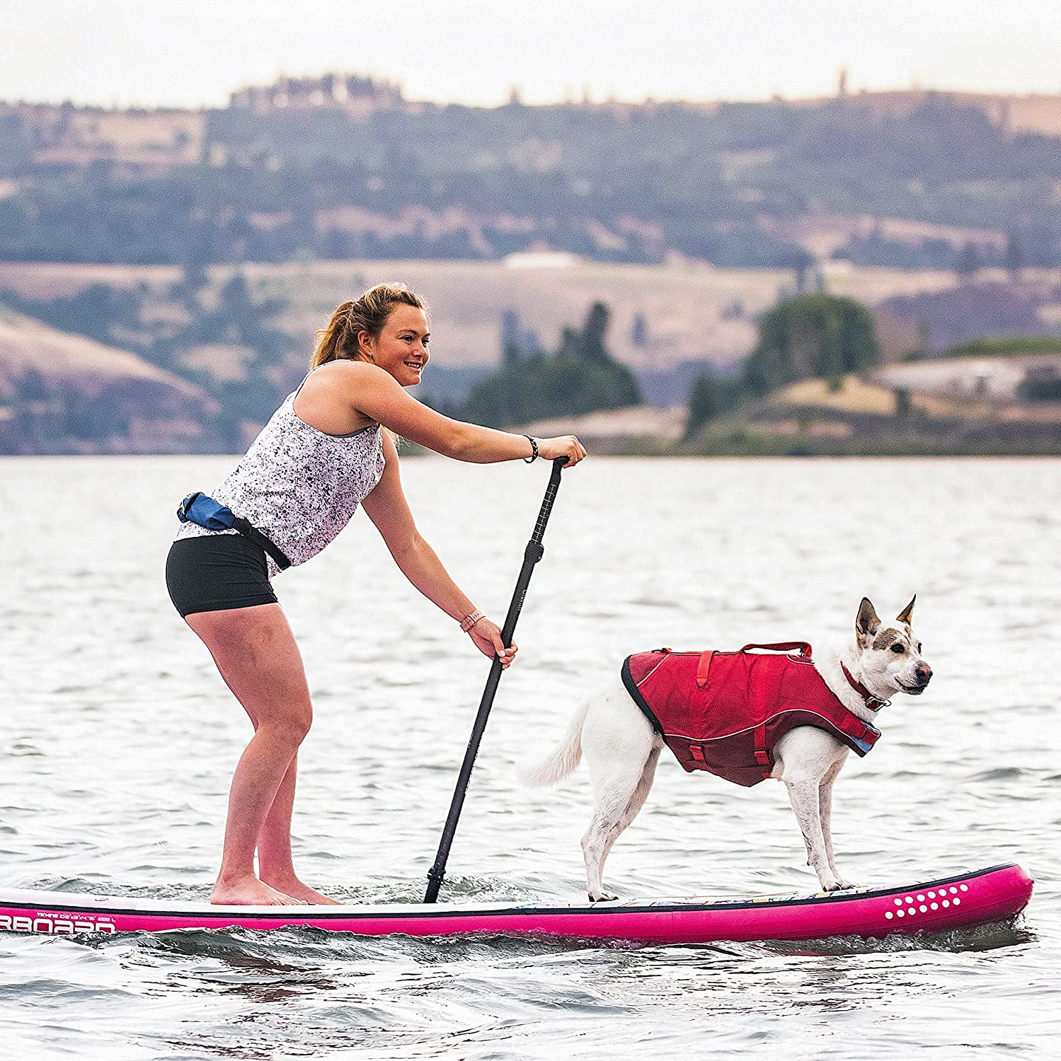 Hund mit roter Kurgo Schwimmweste steht auf einem SUP-Board neben einer Frau auf dem Wasser, ideal für Wassersport und gemeinsame Abenteuer.